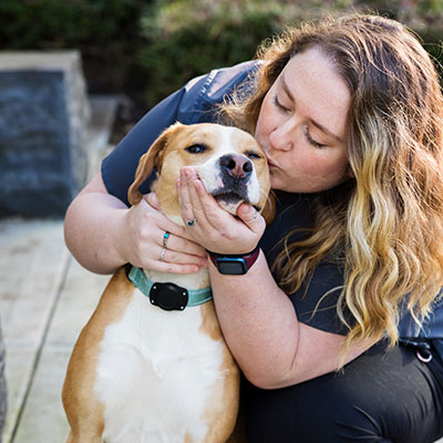 A person with long hair, wearing a smartwatch, is gently embracing and kissing a brown and white dog outdoors. The dog is wearing a light-colored collar and looks content. The background features greenery and stone steps.