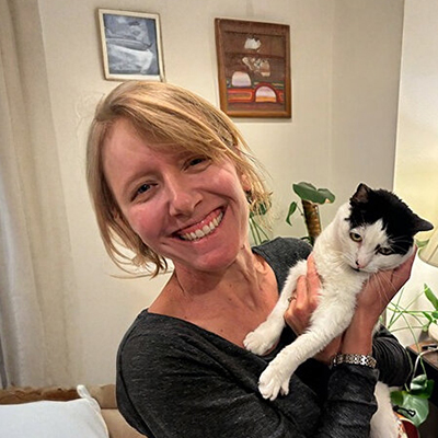 A person with short blonde hair smiles while holding a black and white cat indoors. The room contains framed artwork and green plants.