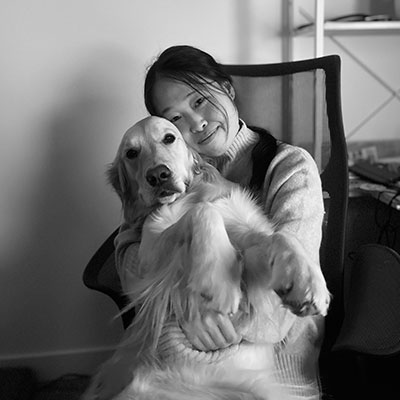 A woman sitting in a chair, smiling while holding a large dog in her lap. The setting is indoors, with a bookshelf in the background. The image is in black and white.